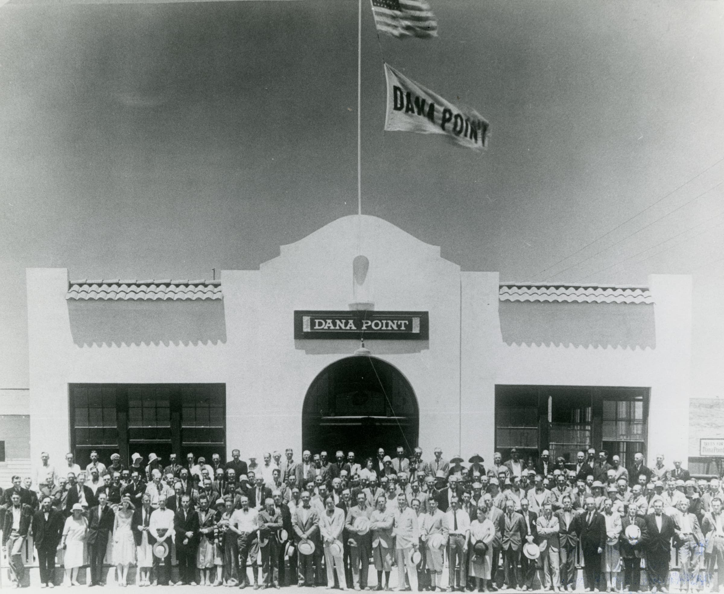 Historic black-and-white photograph of a large crowd in front of a white building with a “Dana Point” sign above its arched doorway and a “Dana Point” pennant flying from the flagpole