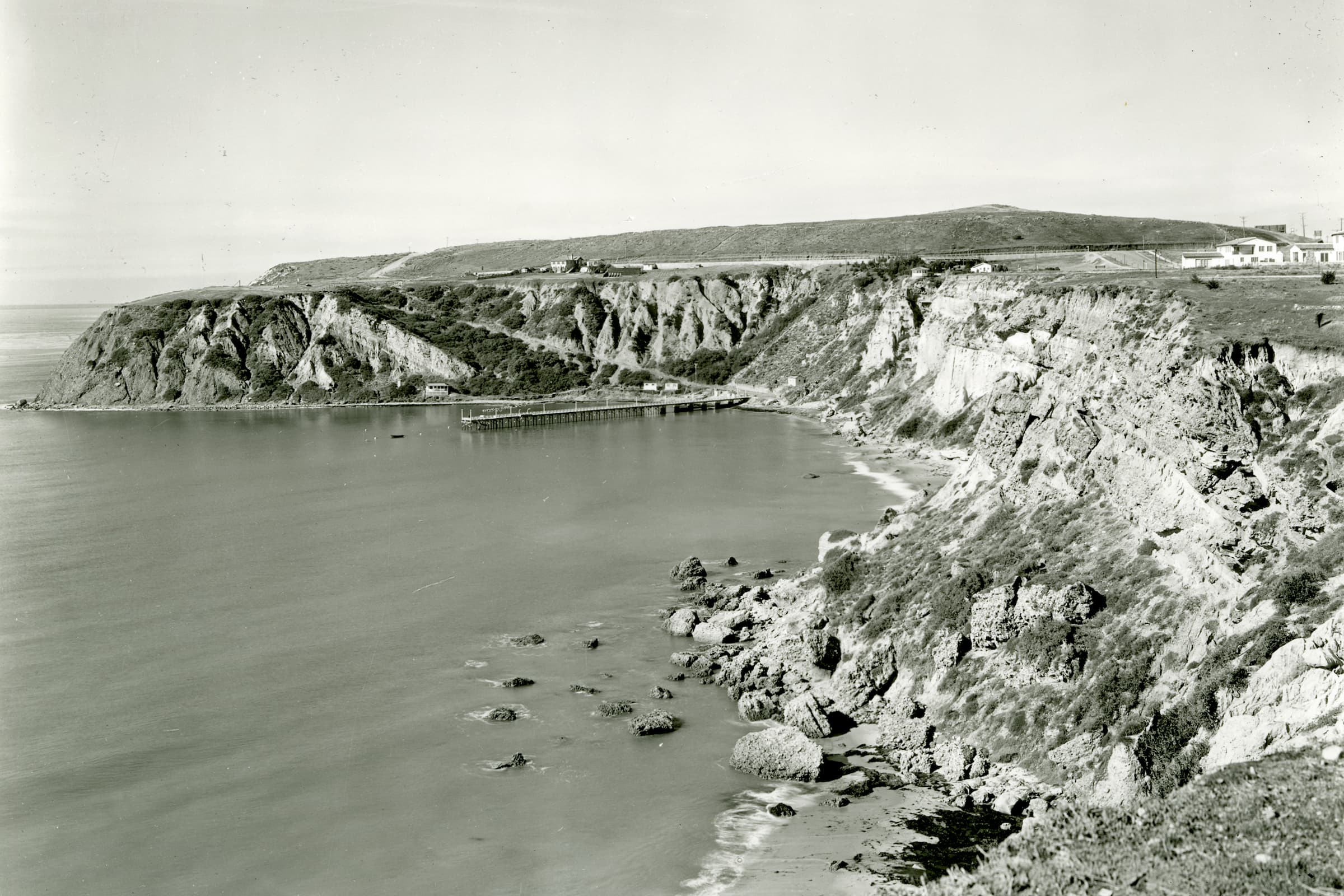 Historic black-and-white photograph of a bay with a wooden pier at the foot of a grassy headland, with eroded cliffs in the foreground